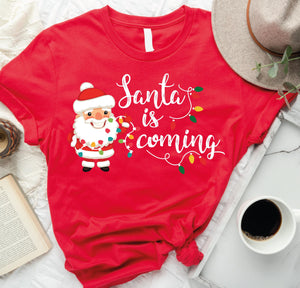 a santa is coming t - shirt on a bed next to a cup of coffee